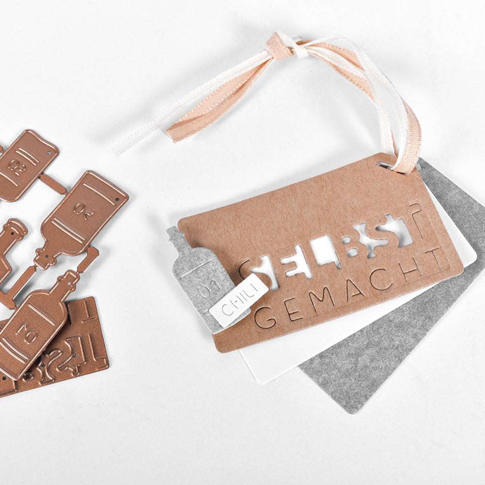 Braune und graue Alexandra Renke-Anhänger mit ausgeschnittenen Buchstaben, Bändern und kleinen Metallstanzteilen sind auf weißem Hintergrund angeordnet.