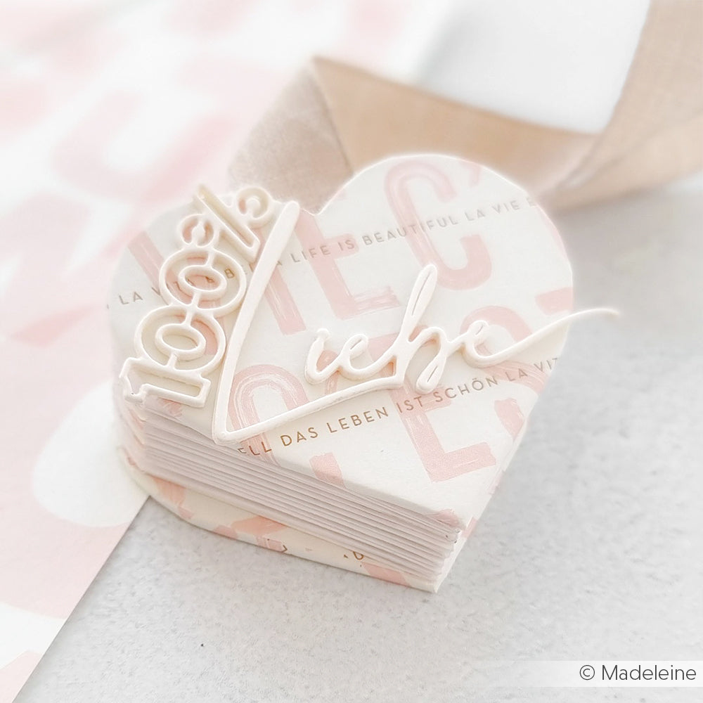 Herzförmiger Papierstapel von Alexandra Renke mit "100% Liebe" in Schreibschrift, präzise gefertigt mit der Motivstanze. Präsentiert auf einem pastellfarbenen Hintergrund - ideal für kreative Projekte mit Designpapieren.