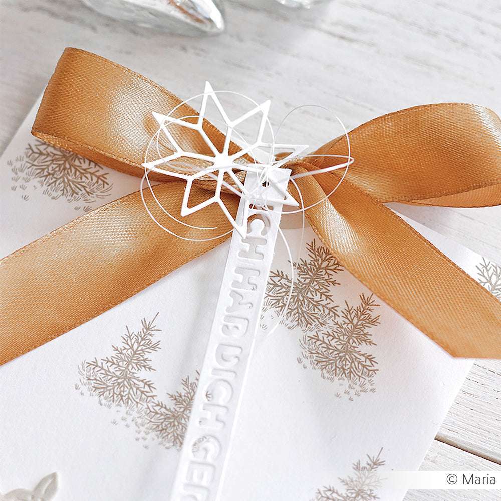 Weiße Karte von Alexandra Renke, hergestellt mit hochwertigen Designpapieren, mit goldenem Band und detaillierter weißer Sternverzierung von DieCut Systeme. Ausgestellt auf einer hellen Holzoberfläche.