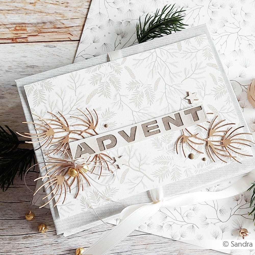 Alexandra Renke Weiße Adventskarte mit goldenen Akzenten, Blattmotiv und dekorativen Sternen aus Motivstanze Adventszahlen, auf einer Holzfläche mit Grünzeug präsentiert.