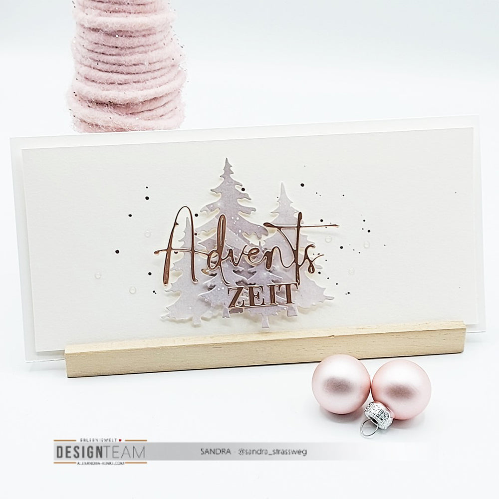 Eine handgefertigte Karte von Alexandra Renke mit dem Motiv "Adventszeit" über rosa Bäumen, hergestellt mit Motivstanze Adventszauber und Designpapier, präsentiert neben rosa Ornamenten und einem rosa Weihnachtsbaum.