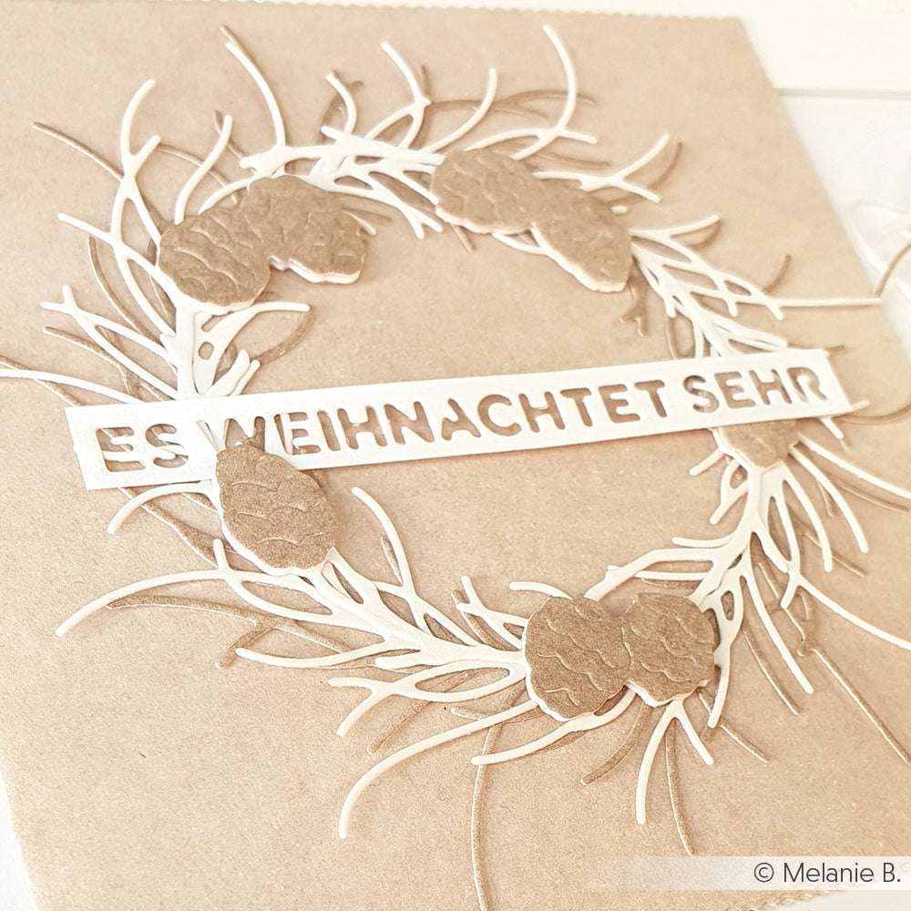Ein mit Alexandra Renke Motivstanze und Designpapieren gebastelter Kranz aus Tannenzapfen mit dem Text ES WEIHNACHTET SEHR auf braunem Hintergrund.