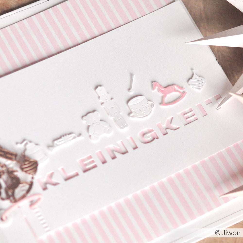 Eine Karte von Alexandra Renke zeigt "KLEINIGKEIT" und kleine weiße Spielzeugformen, die mit der Motivstanze auf einem rosa-weiß gestreiften Hintergrund gestaltet wurden.