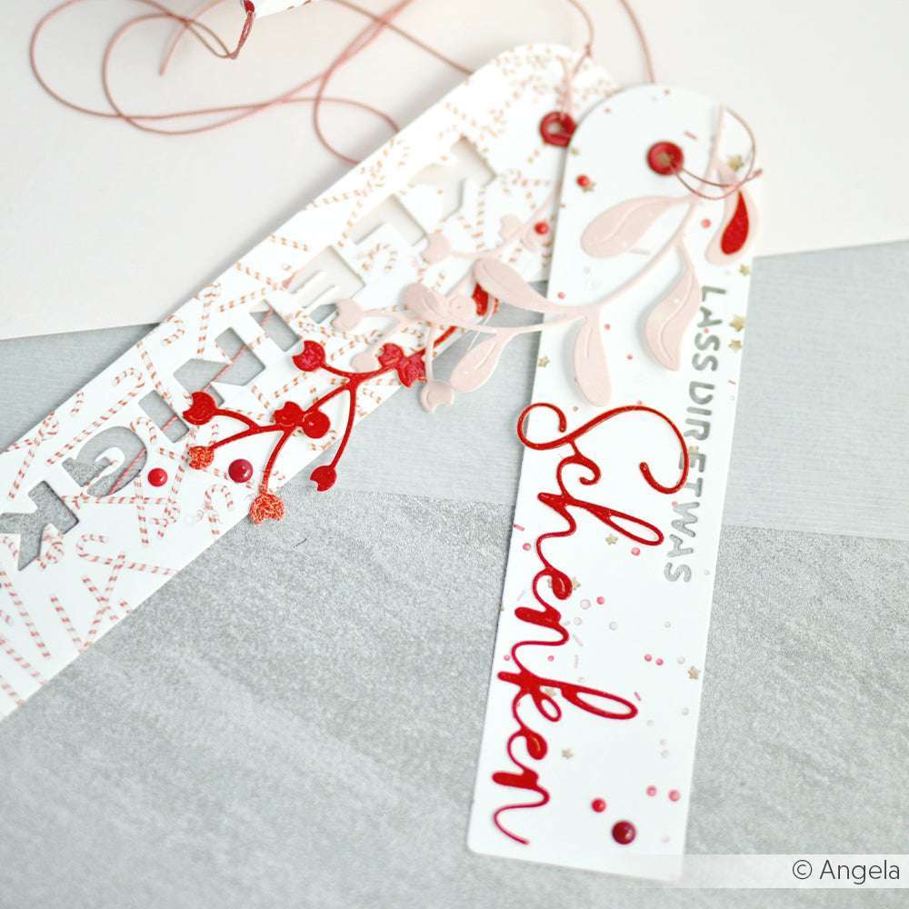 Zwei dekorative Lesezeichen von Alexandra Renke mit roten und rosafarbenen Motiven auf hochwertigen Designpapieren - eines davon mit dem Wort "Schenken" - liegen auf einem Tisch.