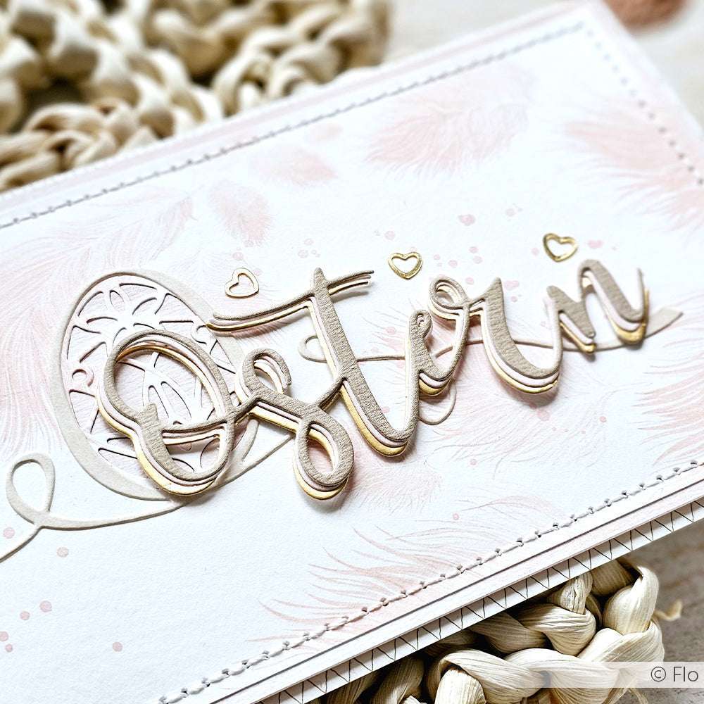 Eine dekorative Karte von Alexandra Renke mit dem Schriftzug "Ostern" in goldener Schrift und pastellrosa Federmotiven, hergestellt mit der Motivstanze Frohe Ostern und hochwertigen Designpapieren für einen festlichen Touch.