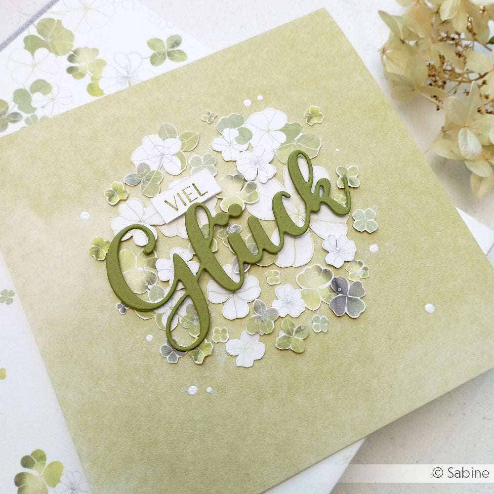 Eine grüne Alexandra Renke Grußkarte mit "Viel Glück" und Kleemotiven, hergestellt mit dem Motivstanze Schriften-Duo Kleines Glück und DieCut Systeme, umgeben von Trockenblumen.