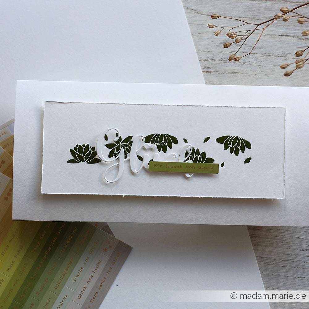 Eine handgefertigte weiße Karte mit Blumenausschnitten, die mit Alexandra Renke Dies hergestellt wurde und mit einem grünen Etikett mit der Aufschrift Ein Hauch von Glück versehen ist.