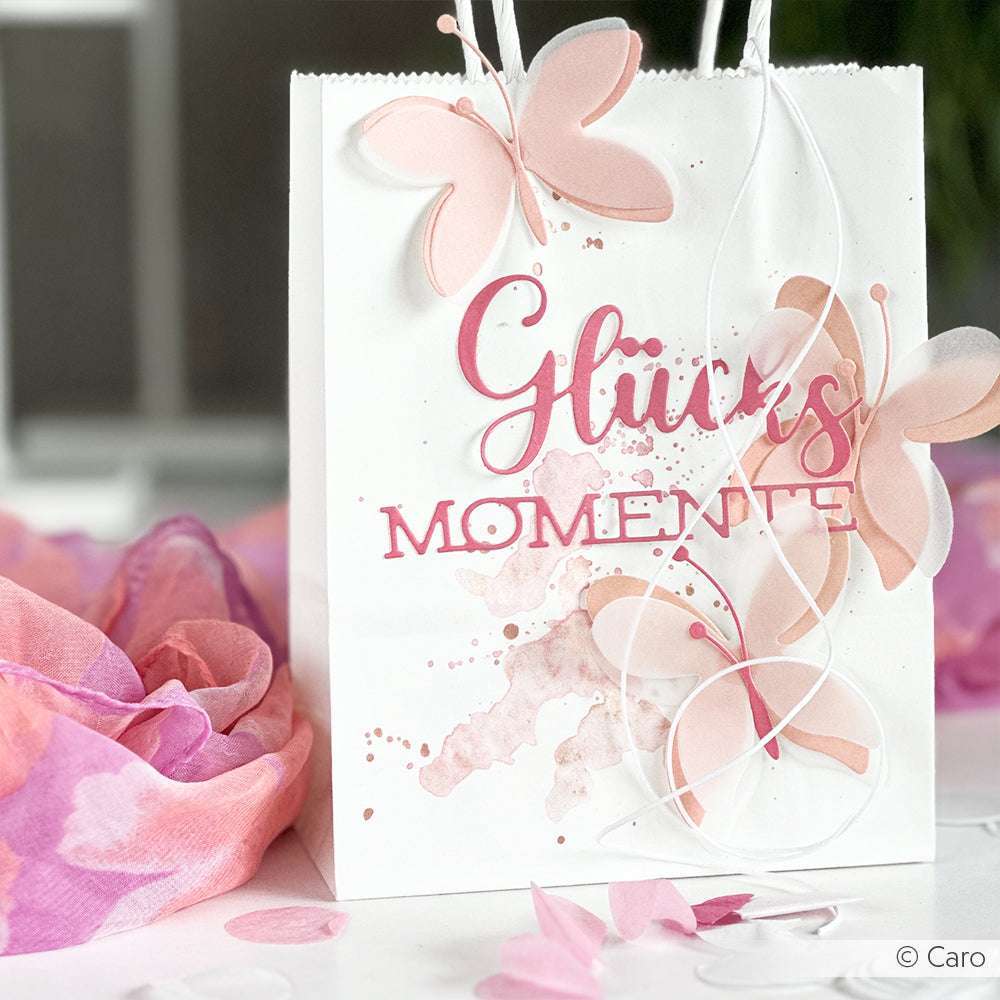 Weiße Geschenktüte mit rosa Schmetterlingen und dem Schriftzug "Glücks Momente", gestaltet mit Alexandra Renke Designpapieren und akzentuiert mit dem Motivstanze Schriften-Duo Glücksmomente. Umgeben von rosa und lila Stoff.