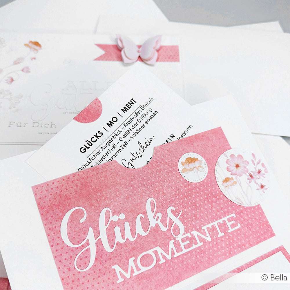 Rosa-weiße Glücksmomente-Karte mit Blumen- und Schmetterlingsdetails, gestaltet mit Alexandra Renke Designpapieren und dem Motivstanze Schriften-Duo Glücksmomente, umgeben von passenden Briefumschlägen.