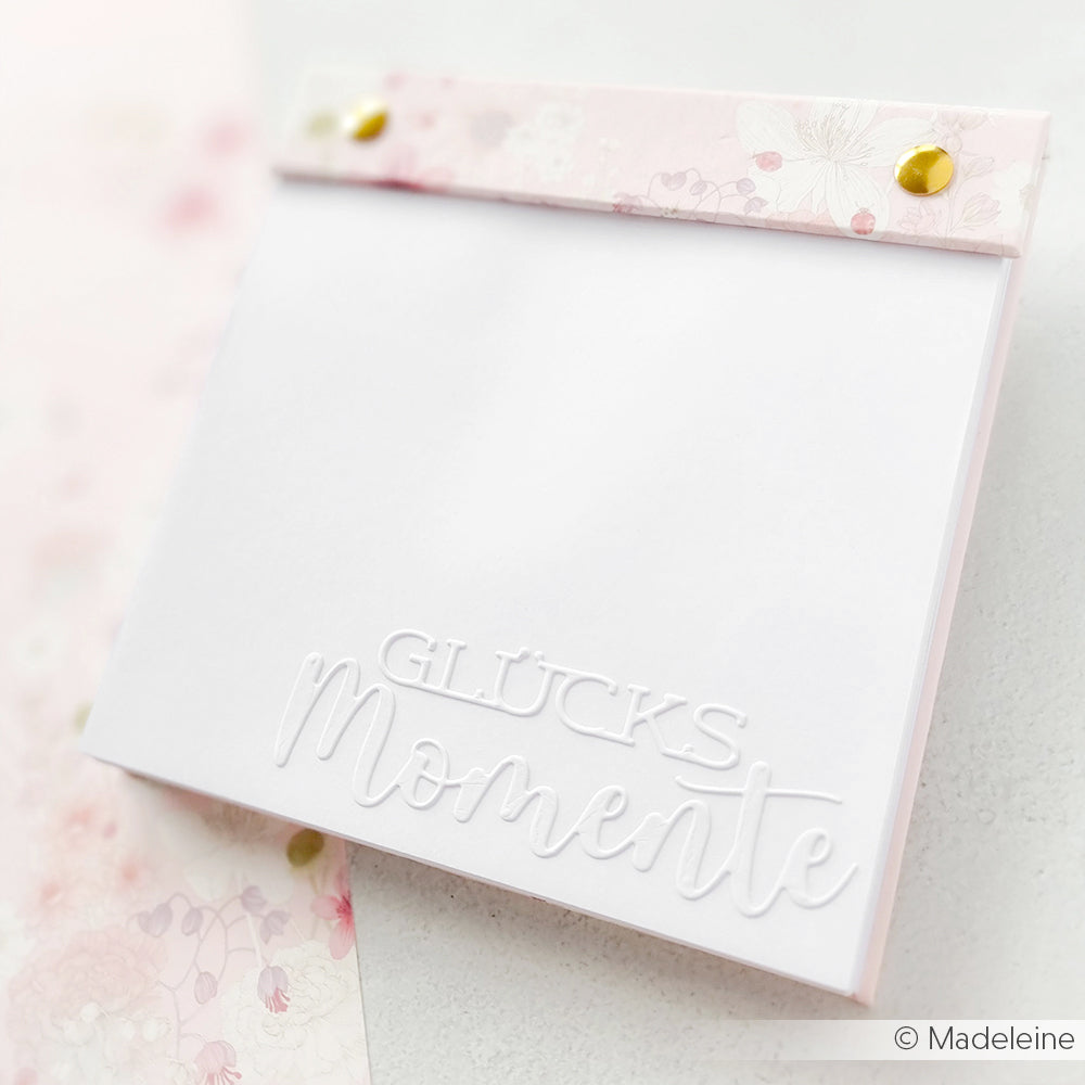 Ein Notizblock von Alexandra Renke mit Glücksmomente-Prägung auf dem Einband, mit pastellfarbenen Blumenakzenten und goldenen Nieten - ideal zum Kombinieren mit Designpapieren oder Ihrem Lieblingsschriften-Duo Glücksmomente für kreative Projekte.