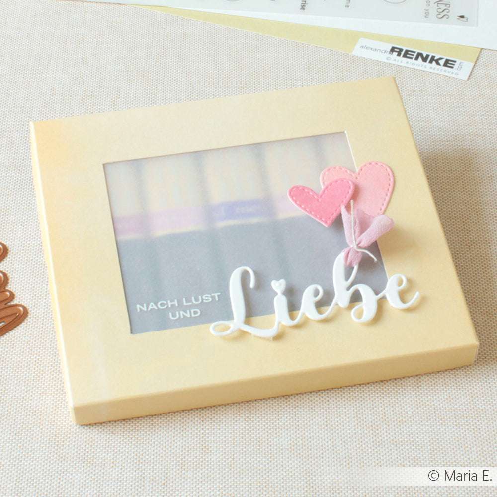 Eine beigefarbene Geschenkbox mit Klarsichtfenster, rosafarbenen Herzchen und dem Schriftzug "Liebe" auf der Vorderseite, die mit dem Motivstanze Schriften-Duo Lieben Dank von Alexandra Renke gestaltet wurde und einen charmanten Touch hat.