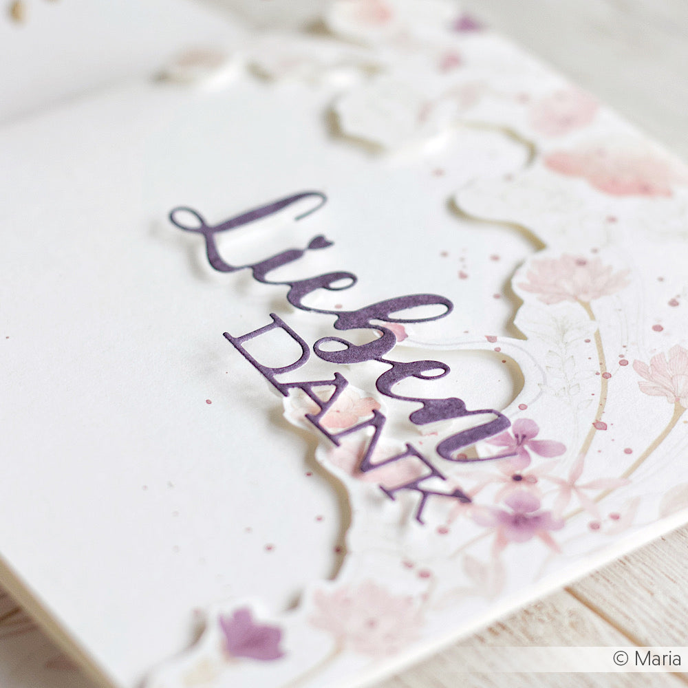 Nahaufnahme einer floralen Dankeskarte, die mit Alexandra Renke Schriften-Duo Dies und eleganten Designpapieren gestaltet wurde, mit "Lieben Dank" in lila Schrift auf weißem Papier.