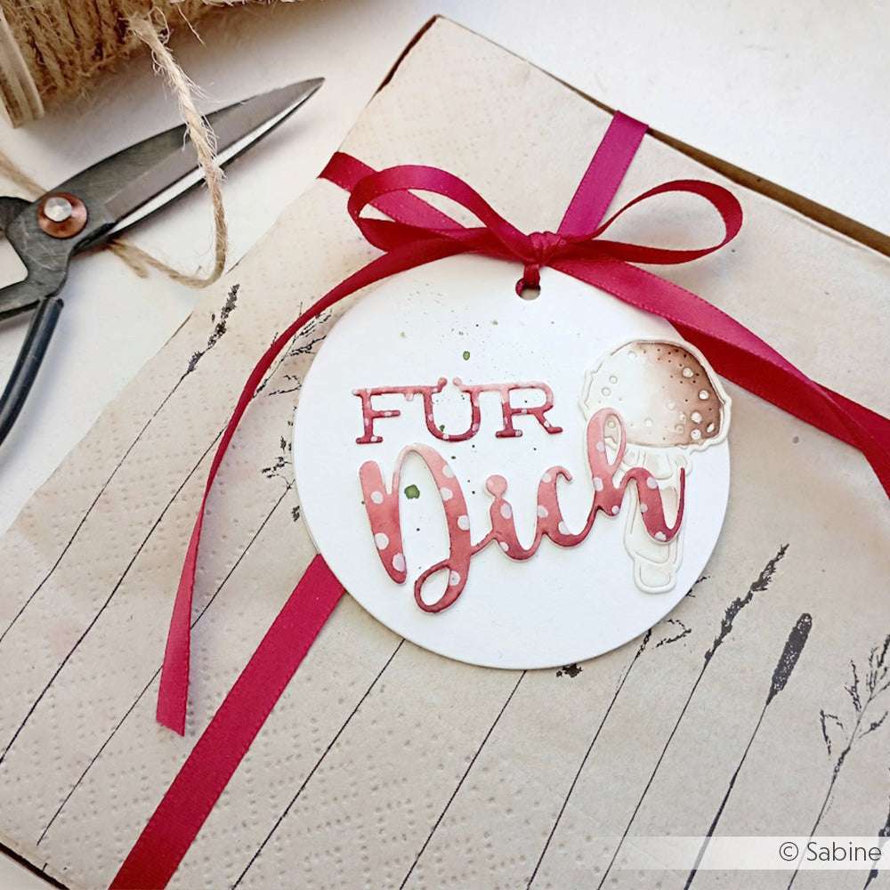 Alexandra Renke runder Geschenkanhänger mit "Für Dich" in roter Schrift und einem Pilz, angefertigt mit dem Motivstanze Schriften-Duo, gebunden mit einem roten Band auf einem eingepackten Geschenk.
