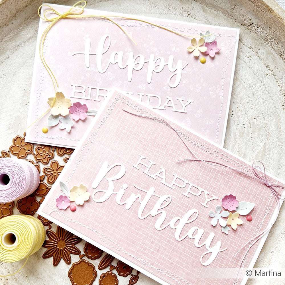 Zwei rosafarbene, handgefertigte Geburtstagskarten von Alexandra Renke mit Blumendekorationen, Bindfäden und einer eleganten Schrift mit einer Motivstanze Happy Birthday, die auf einer Holzfläche präsentiert wird.