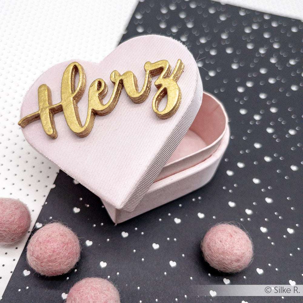 Eine rosafarbene herzförmige Schachtel mit der Aufschrift "Herz" aus Alexandra Renke Designpapieren 200g steht zwischen rosafarbenen Filzkugeln auf einem gepunkteten Hintergrund.