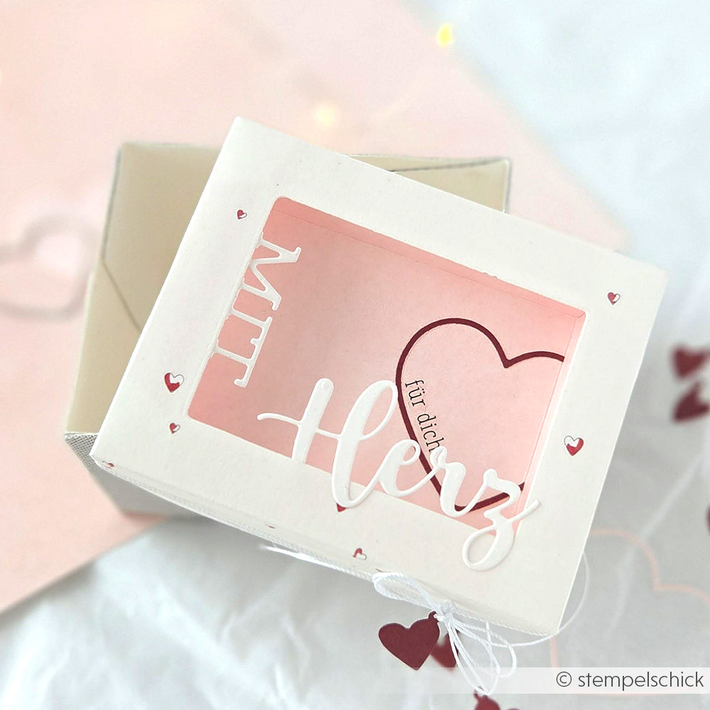 Eine weiße Alexandra Renke Geschenkbox mit Herzausschnitten und "Mit Herz" auf dem Deckel, hergestellt mit Stanzsystemen, enthält eine rosa Karte mit Herzmotiv.