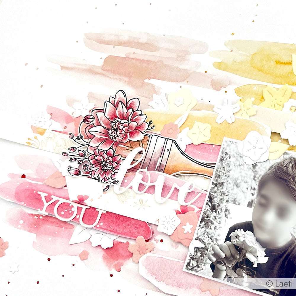 Eine Person hält eine Blume mit "Love You"-Text und Blumenkunst auf einem rosa-gelben Aquarell-Hintergrund. Ausgestattet mit Alexandra Renke Designpapieren und dem Motivstanze Schriften-Duo Love You für einen handwerklichen Touch.