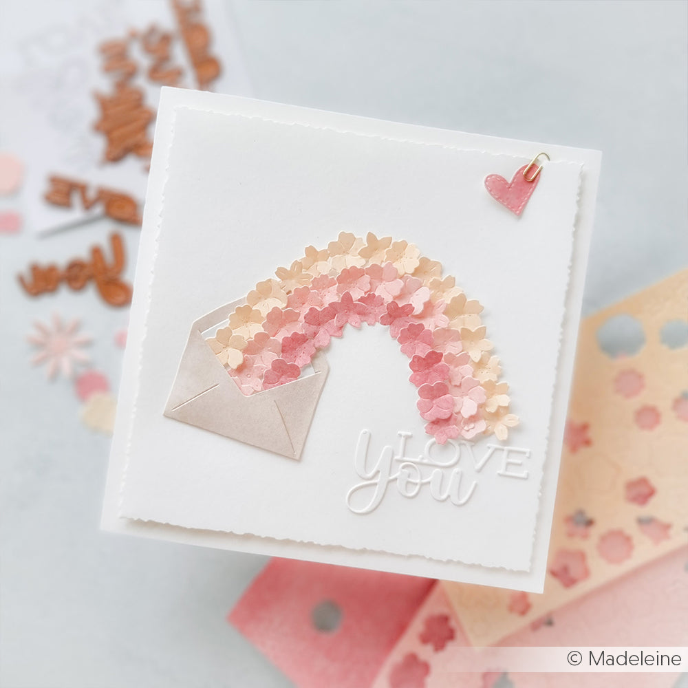 Eine handgefertigte Karte von Alexandra Renke mit einem Umschlag, pastellfarbenen Regenbogenherzen, dem Text "Love You" und einem kleinen Herzanhänger - erstellt mit den Motivstanze Schriften-Duo Love You Dies.