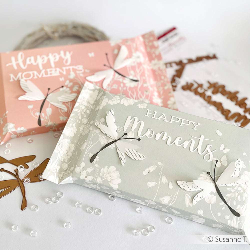 Zwei dekorative Geschenkboxen mit Happy Moments Text und Libellenmotiven von Alexandra Renke, umgeben von Designpapieren und Bastelmaterial - perfekt für die Verwendung mit Motivstanze Schriften-Duo oder DieCut Systemen.