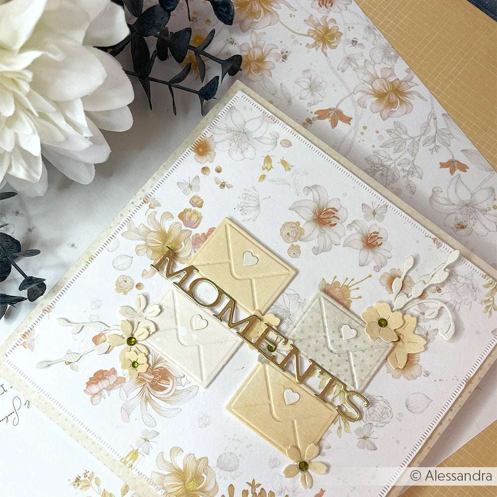 Eine Blumenkarte von Alexandra Renke mit Designpapieren, pastellfarbenen Umschlägen, Papierblumen und "MOMENTS" in goldener Schriften-Duo Happy Moments-Schrift auf der Vorderseite.