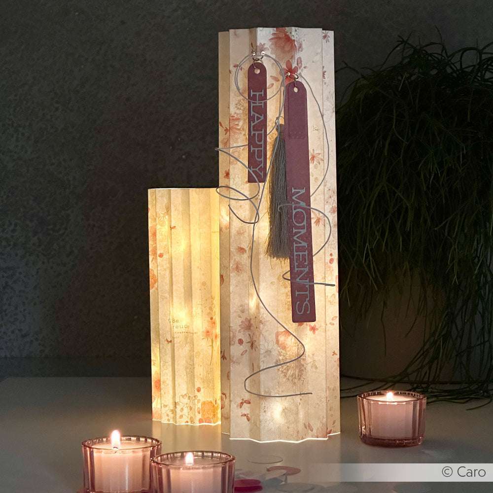 Zwei dekorative Papierlaternen aus Alexandra Renke Designpapieren mit sanftem Licht leuchten neben drei brennenden Kerzen auf einer weißen Fläche und setzen elegante Alexandra Renke DieCut Systeme Akzente.