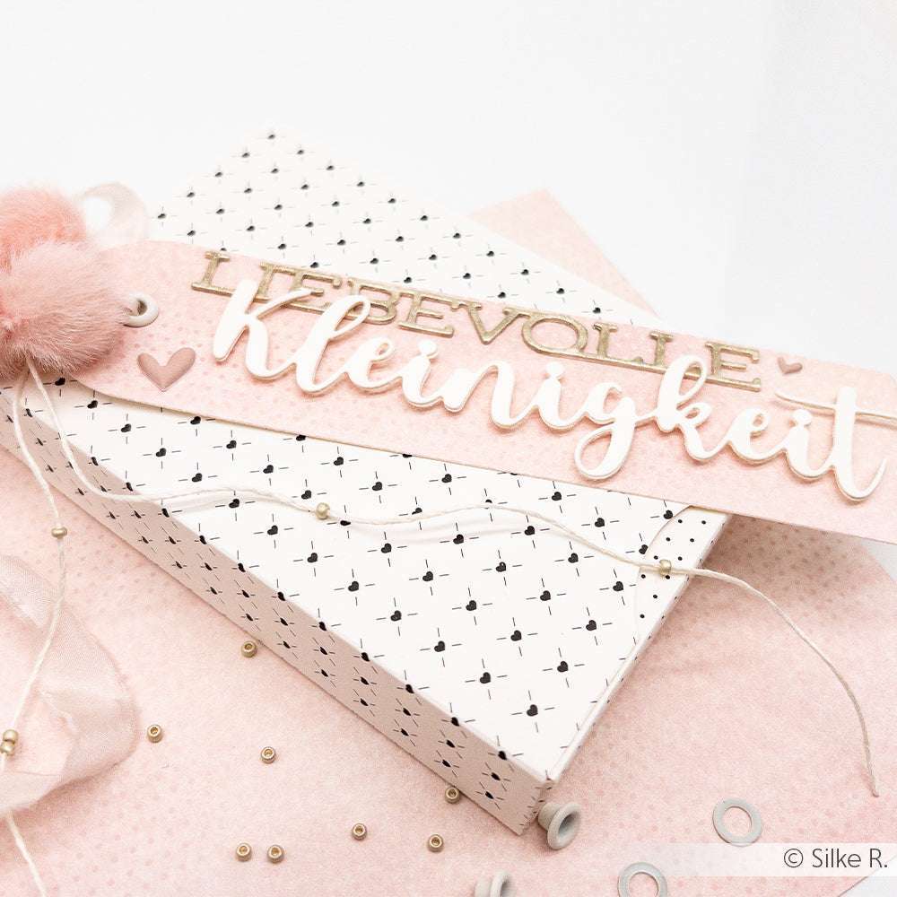 Alexandra Renke Geschenkbox mit rosa "LIEBEVOLLE Kleinigkeit"-Anhänger, verziert mit einem Pompon und aus Motivstanze, auf gepunktetem Hintergrund.