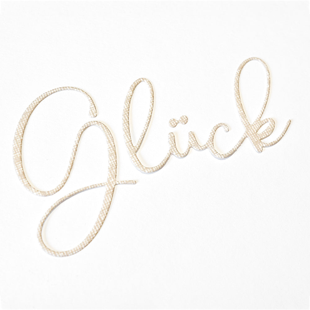 Das Wort Glück, gestaltet mit der Alexandra Renke Motivstanze Glück Schreibschrift, steht in eleganter, strukturierter Schreibschrift auf hochwertigen Designpapieren vor einem strahlend weißen Hintergrund.