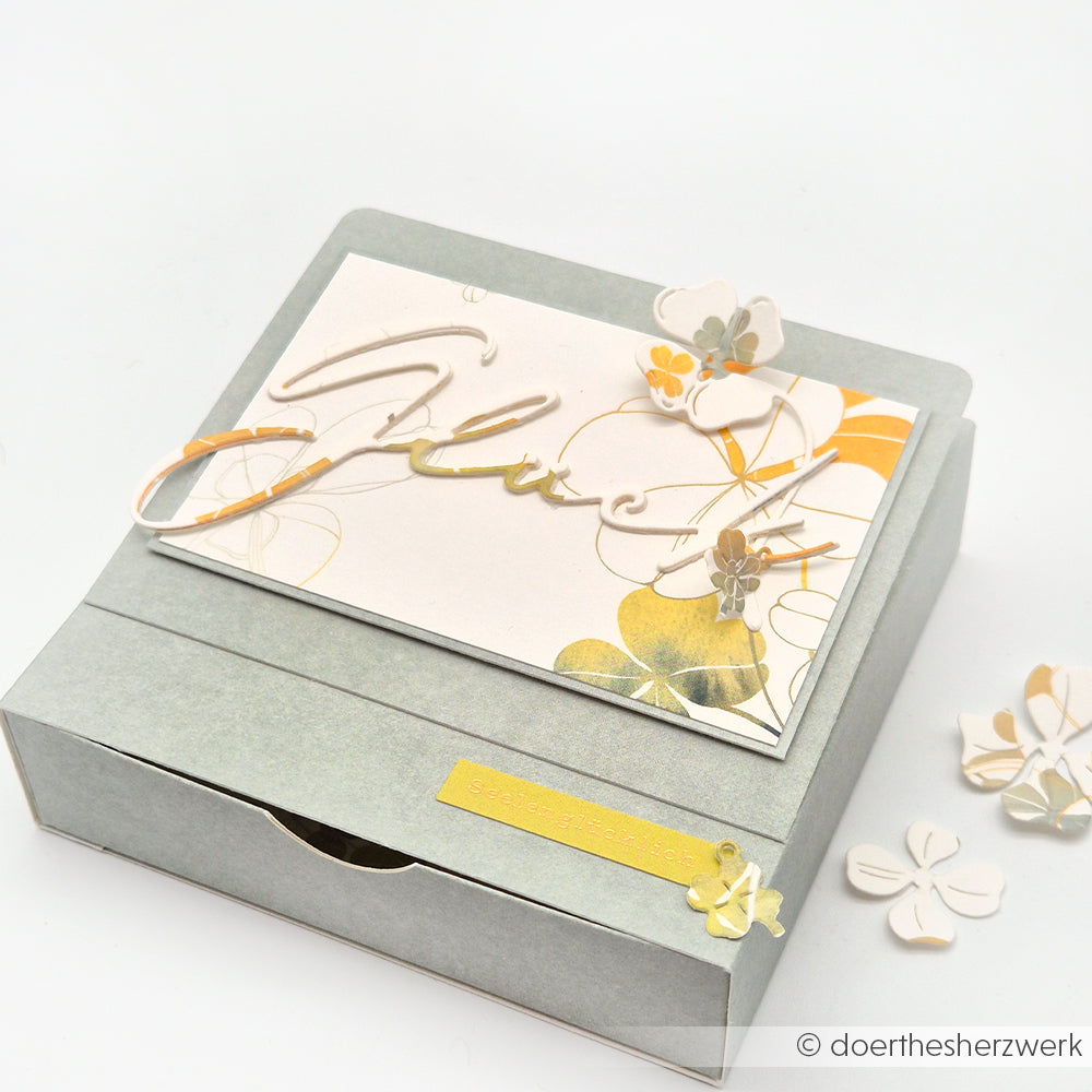 Eine graue Geschenkschachtel mit einer Blumenkarte, die mit kleinen Papierblumen aus der Motivstanze Glück von Alexandra Renke verziert ist und mit eleganten Designpapieren verziert wurde.
