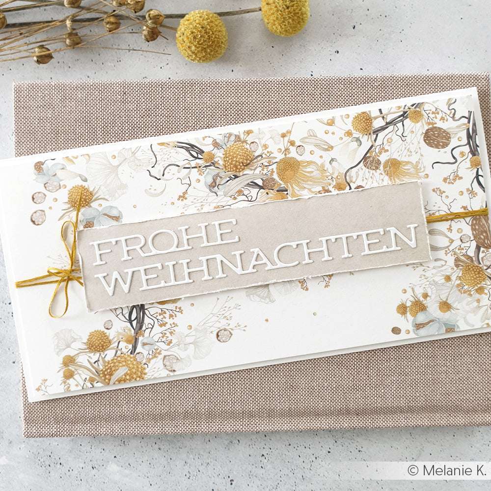 Eine festliche Karte von Alexandra Renke mit "Frohe Weihnachten" in Weiß, gestaltet mit dem Motivstanze Schriften-Duo, auf einem floralen Hintergrund mit eleganten Goldakzenten.
