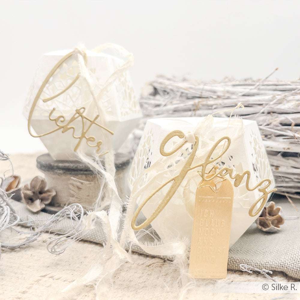 Zwei weiße Laternen mit goldenen Schriftzügen aus dem Alexandra Renke Motivstanze Wörterset, rustikaler Schnur, Tannenzapfen und Treibholz sind kunstvoll auf einem neutralen Hintergrund arrangiert.