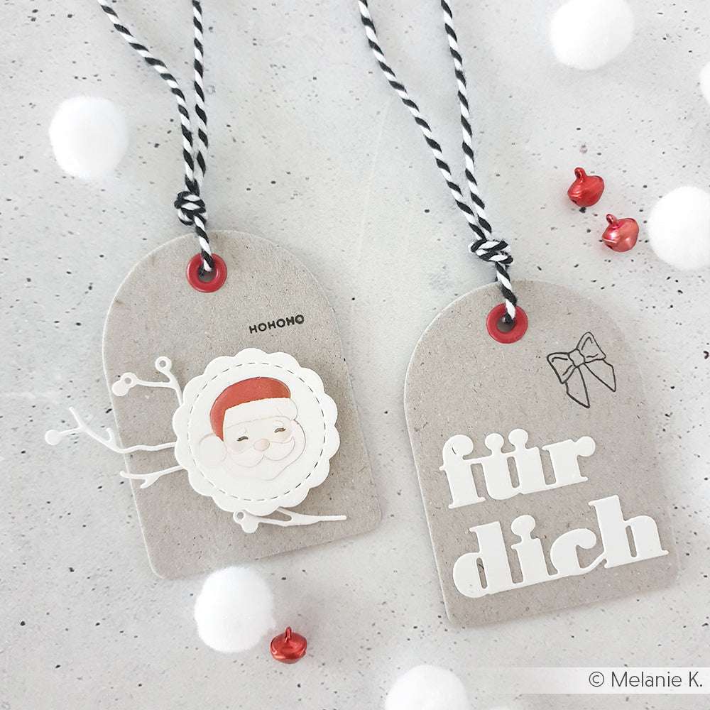 Zwei Geschenkanhänger aus Festtagspapier von Alexandra Renke - einer mit einem Weihnachtsmanngesicht und "hohoho", der andere mit "für dich" und einer kleinen Schleife - sind auf einer grauen Fläche ausgestellt.
