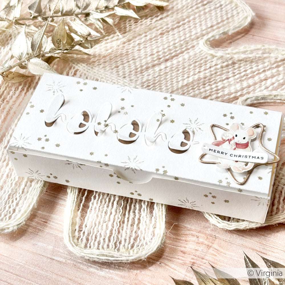 Weiße, weihnachtliche Geschenkbox von Alexandra Renke mit einem "Hohoho"-Ausschnitt, einem Anhänger in Form eines Lebkuchens und aus hochwertigem Designpapier. Ausgestellt auf einer strukturierten Oberfläche.