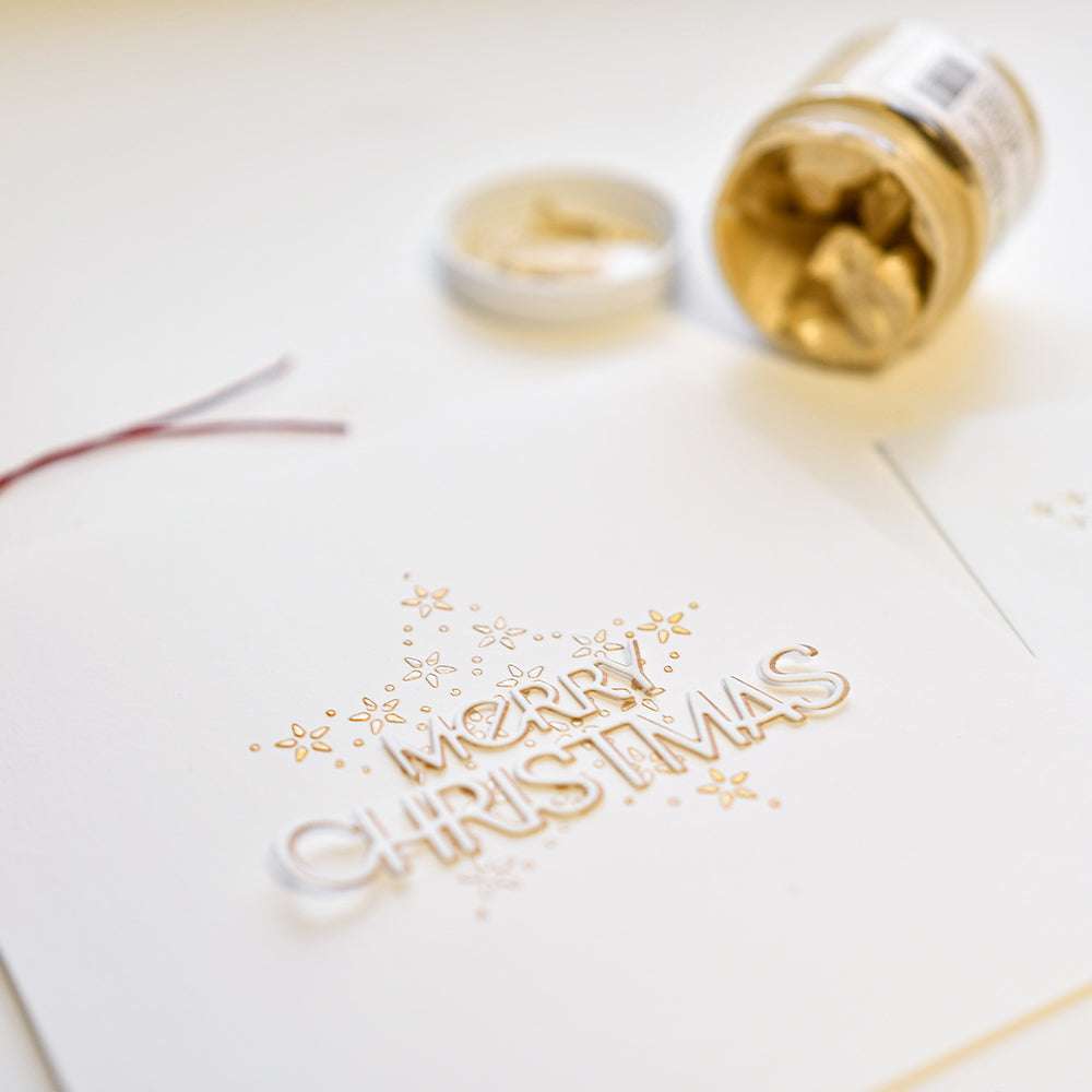 Weiße Karte mit "Merry Christmas", ausgestanzt mit der Alexandra Renke Motivstanze Merry Christmas, verziert mit goldenen Sternen, neben einem offenen Glas mit goldfarbenen Teilen.