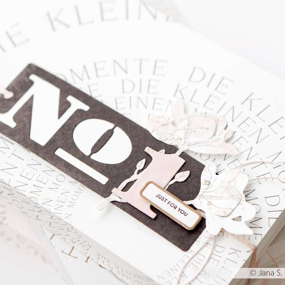 Ein dekorativer "Noel"-Anhänger von Alexandra Renke, gefertigt mit Deep Edge Motivstanzen, mit floralen Akzenten und einem kleinen "Just for You"-Etikett auf weißem, textgemustertem Hintergrund.