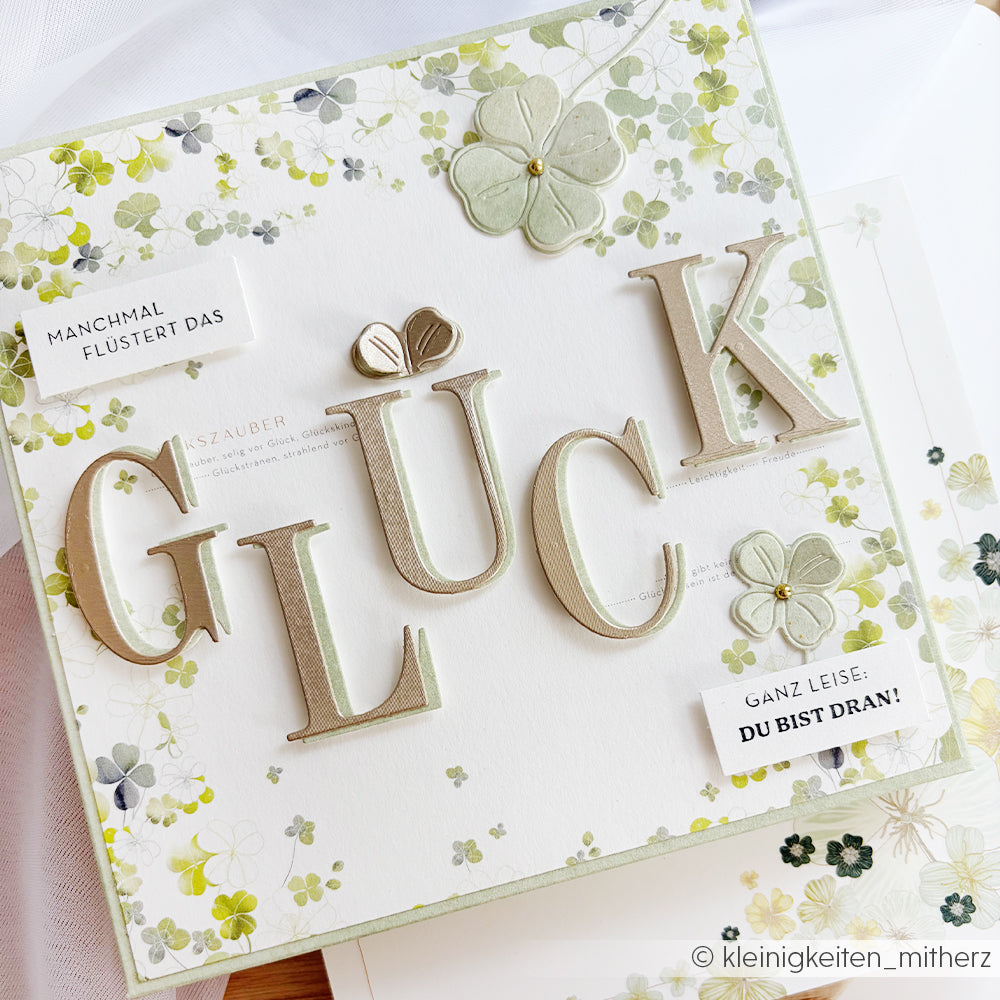 Quadratische Karte mit fettgedrucktem "GLÜCK", hergestellt mit Alexandra Renkes Motivstanze Classy Alphabet, mit floralen Akzenten und deutschem Text auf weißem und grünem Hintergrund.