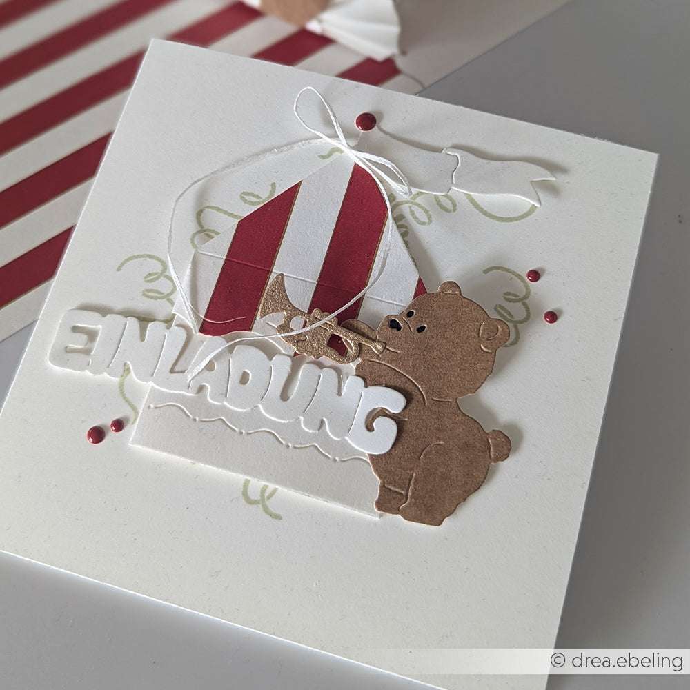 Handgemachte Einladungskarte mit einem Bären, einem Zirkuszelt und dem Text "EINLADUNG", verziert mit roten Punkten und einer Schleife. Hergestellt mit Alexandra Renke Produkten für charmante Details.