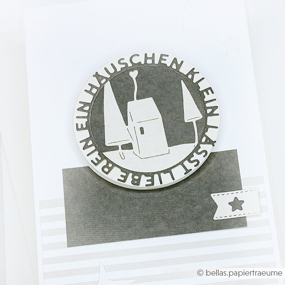 Ein runder Scherenschnitt von Alexandra Renke zeigt ein Haus, Bäume und einen deutschen Text auf einer monochromen Karte, die mit Stern- und Streifendetails verziert ist.