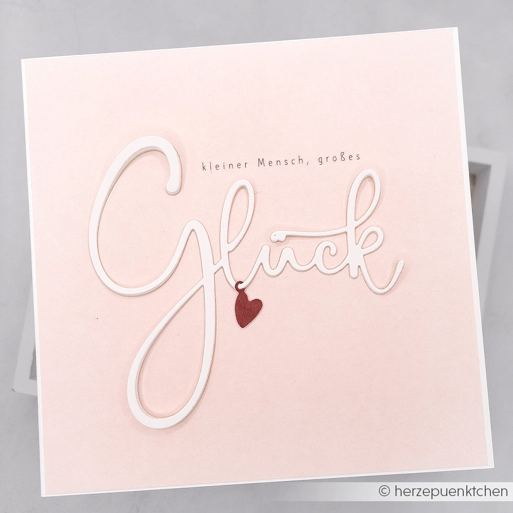 Quadratische, rosafarbene Karte von Alexandra Renke mit dem Spruch "Kleiner Mensch, großes Glück" und einem Herzanhänger auf Glück, gefertigt aus hochwertigen Designpapieren für einen edlen Touch.