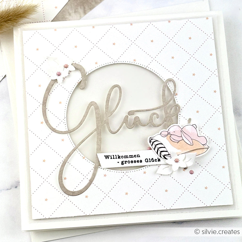 Quadratische Karte von Alexandra Renke mit "Glück" in Schreibschrift, floralen Akzenten und einem deutschen Gruß auf gepunktetem weißem Hintergrund, hergestellt mit DieCut Systemen für präzise Details.