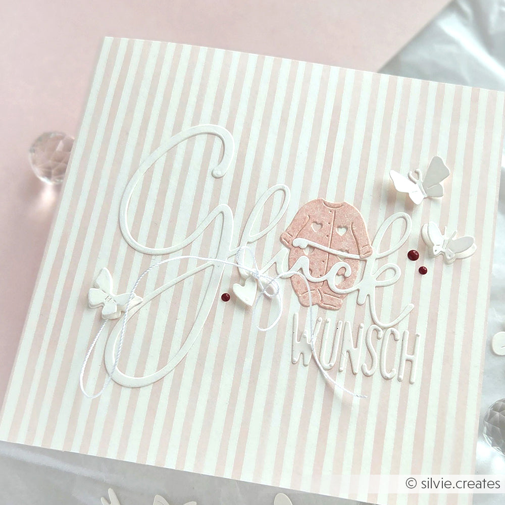 Rosa gestreifte Karte von Alexandra Renke mit "Glückwunsch", weißen Schmetterlingsakzenten und einem rosa Baby-Strampler, der mit dem Motivstanze DieCut System erstellt wurde.