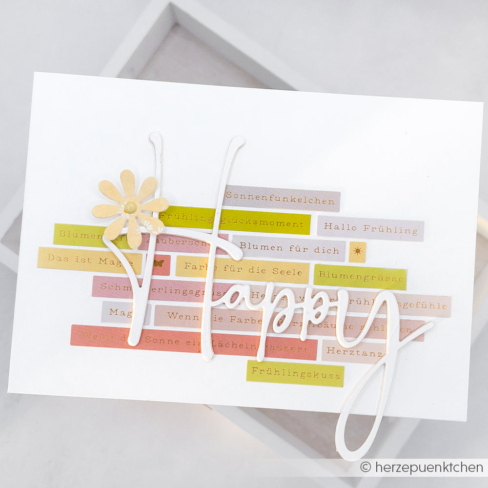 Eine handgefertigte Karte von Alexandra Renke mit farbenfrohen deutschen Textstreifen, einer gelben Blume und "Happy" in fetter Schrift, hergestellt mit hochwertigen Designpapieren für ein einzigartiges und lebendiges Aussehen.