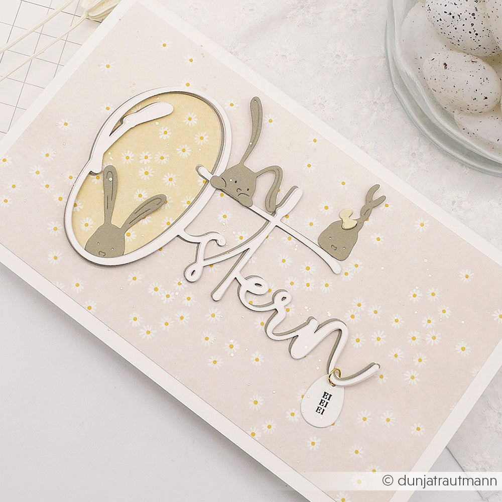 Die Osterkarte von Alexandra Renke zeigt Hasengesichter und -ohren, die "Ostern" bilden, umgeben von weißen und gelben Blumen - ideal für die Verwendung mit der Motivstanze Ostern oder den DieCut Systemen auf Ihren Lieblingsdesignpapieren.