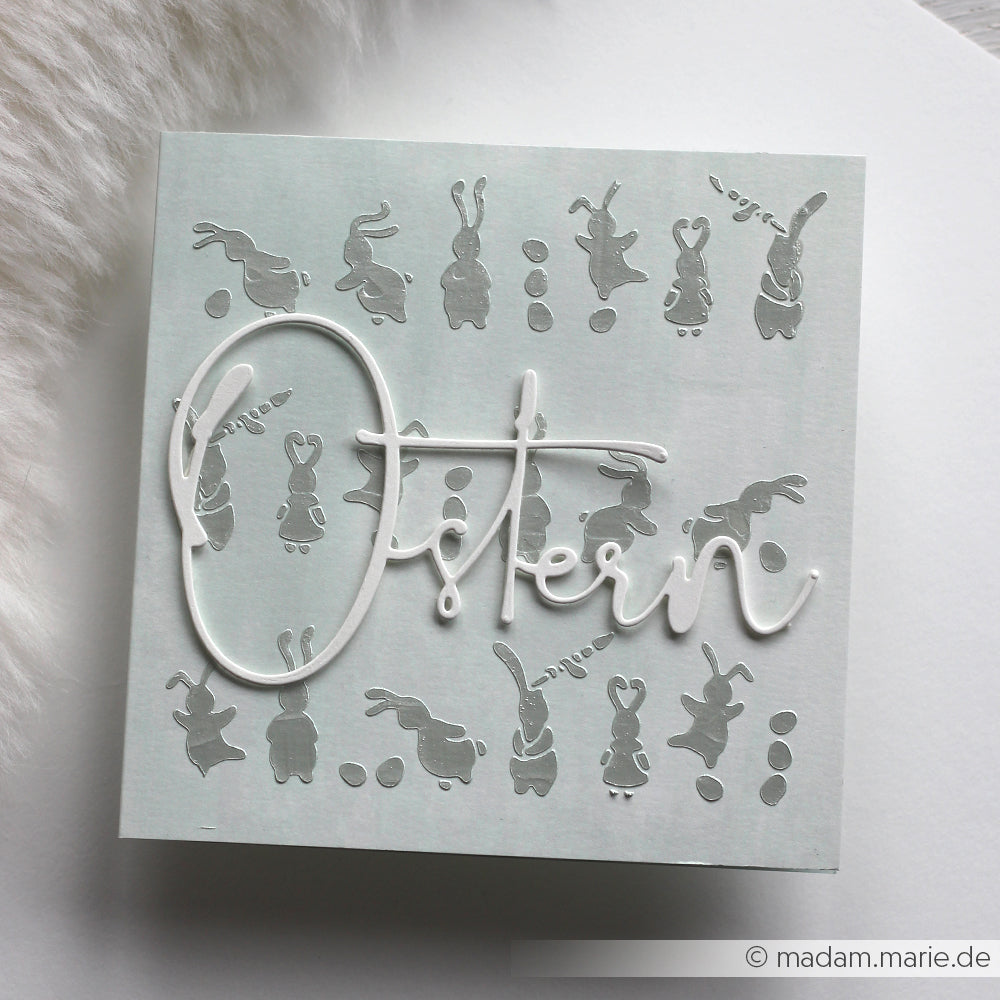 Quadratische Osterkarte von Alexandra Renke aus hochwertigem Designpapier mit Hasen- und Eiersilhouetten, die mit der Motivstanze Ostern ausgestanzt wurden, und dem Schriftzug Ostern" in weißer Schrift auf der Vorderseite.