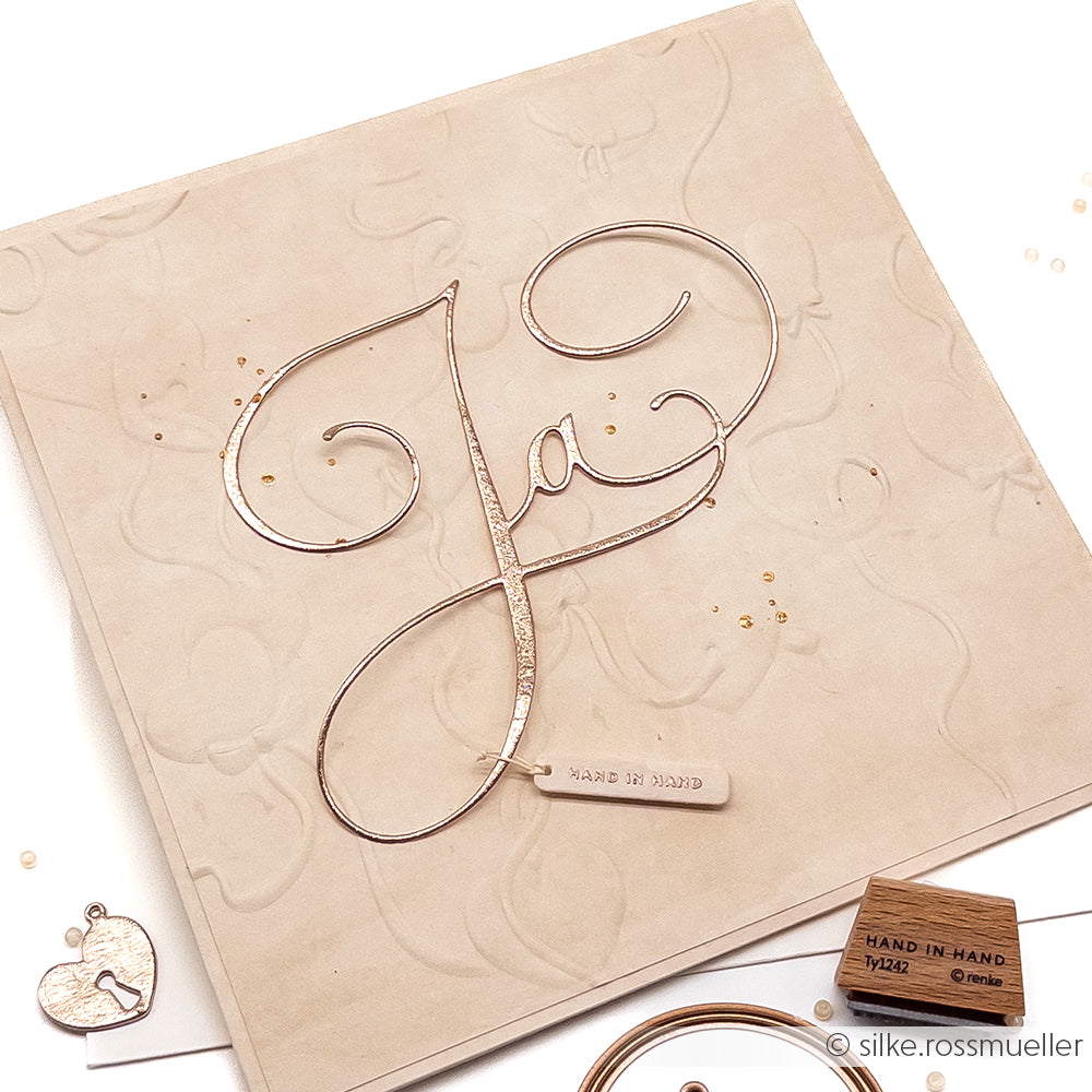 Elegante Karte von Alexandra Renke mit einem kalligrafischen "Ja" aus einer Motivstanze und einem kleinen HAND IN HAND-Anhänger, auf hochwertigem Designpapier mit geprägtem beigem Hintergrund.