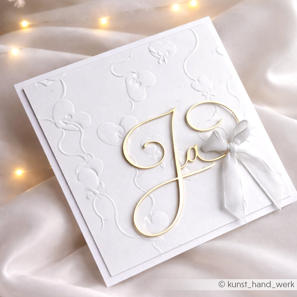 Elegante weiße Hochzeitskarte von Alexandra Renke mit goldener Schrift Ja aus der Motivstanze Kalligrafisch Ja, geprägten Herzen und einer weißen Schleife auf seidigem Designerpapier für einen luxuriösen Touch.