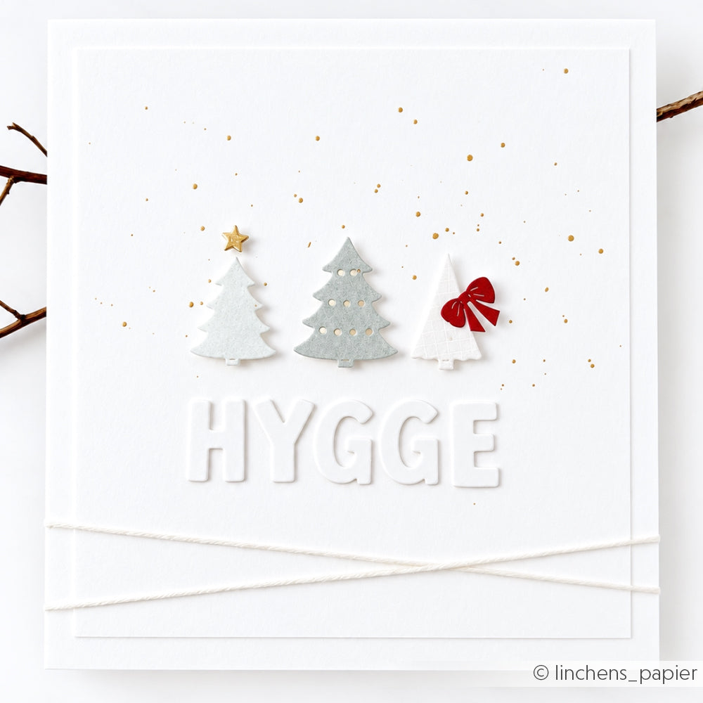 Grußkarte mit drei Weihnachtsbäumen aus Papier, goldenen Glitzerpartikeln und einem Stanzmotiv mit HYGGE-Alphabet.