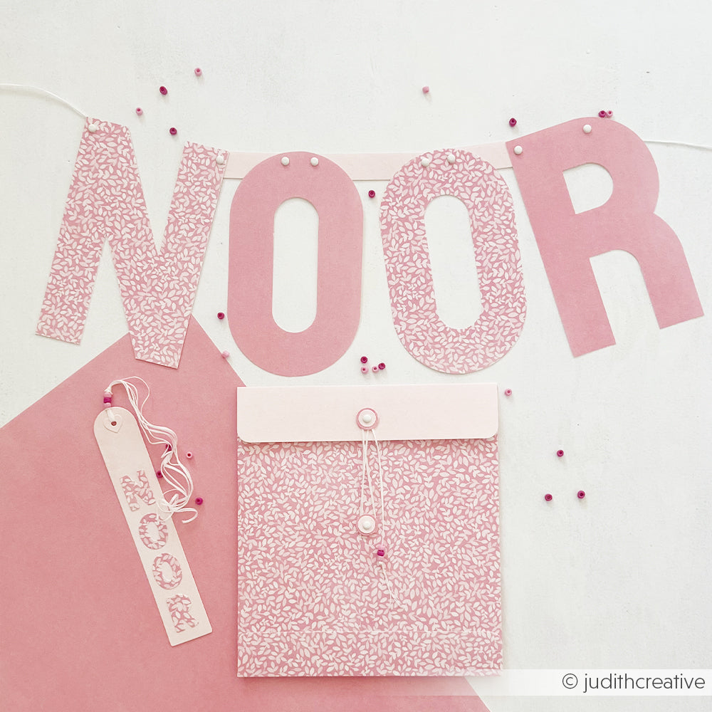 Ein rosa Papierbanner mit der Aufschrift NOOR, ein Lesezeichen und ein Umschlag, die mit den Motivstanze Alphabet-Werkzeugen gestaltet wurden.