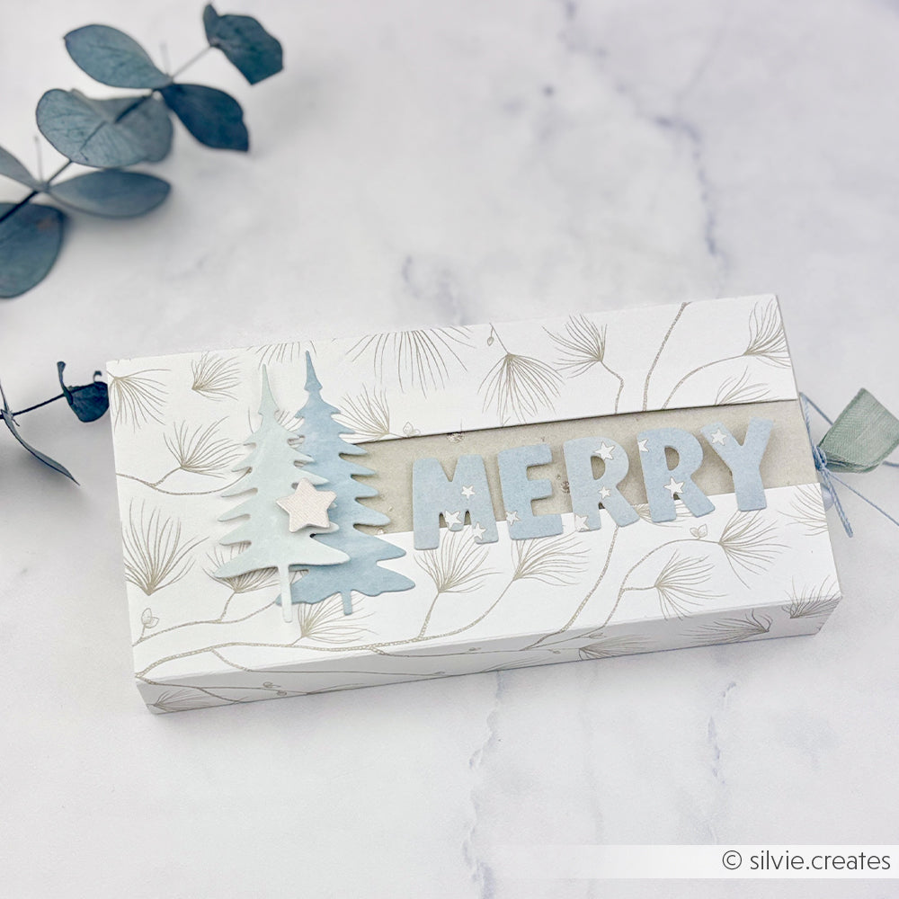 Eine Geschenkbox mit Kiefernmotiven, Papierbäumen und der Aufschrift MERRY, gestaltet mit den Motivstanze Alphabet-Stanzformen.
