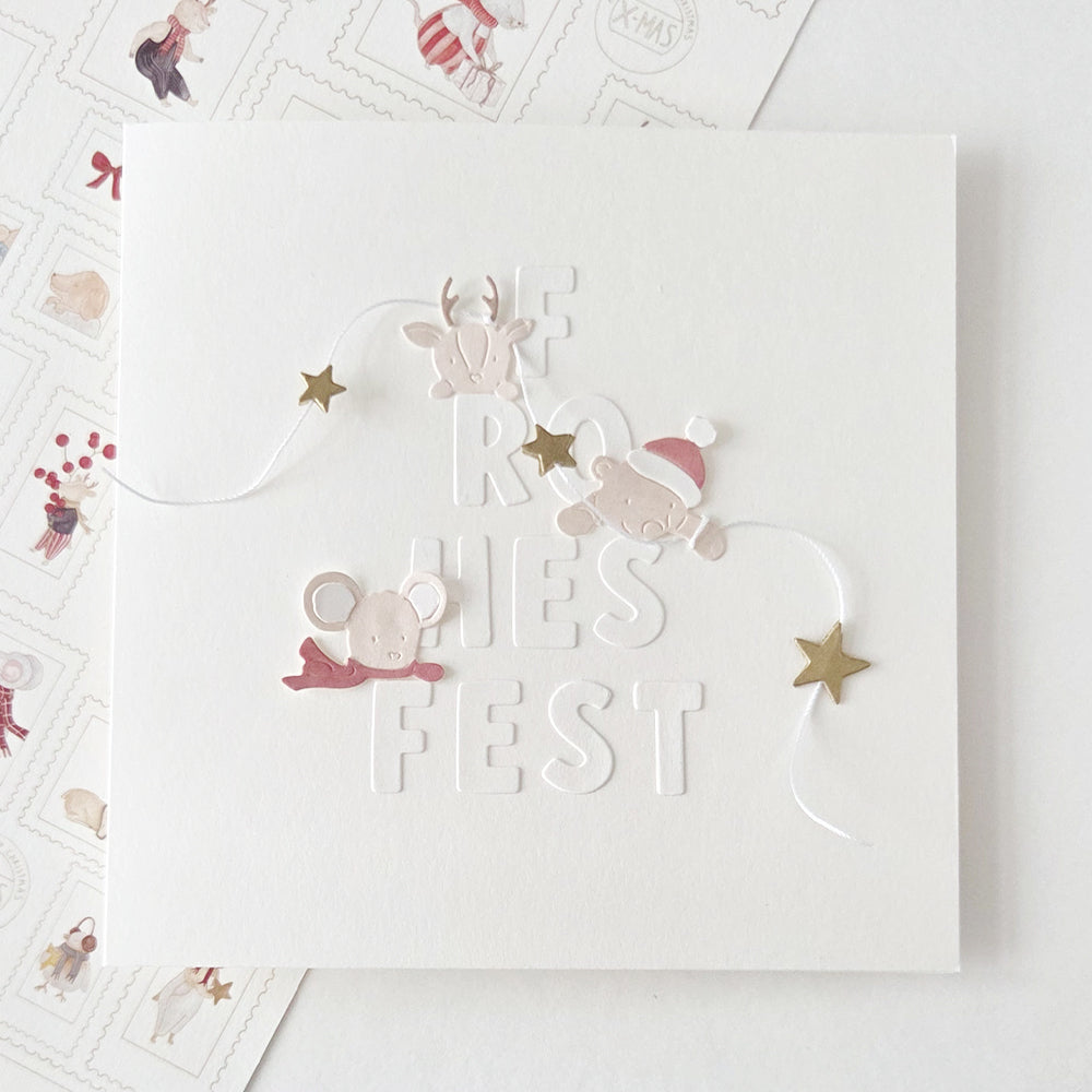 Eine weiße, quadratische Weihnachtskarte mit der Prägung „FROHES FEST“, verziert mit Designpapieren und kleinen Tier- und Sternausschnitten.