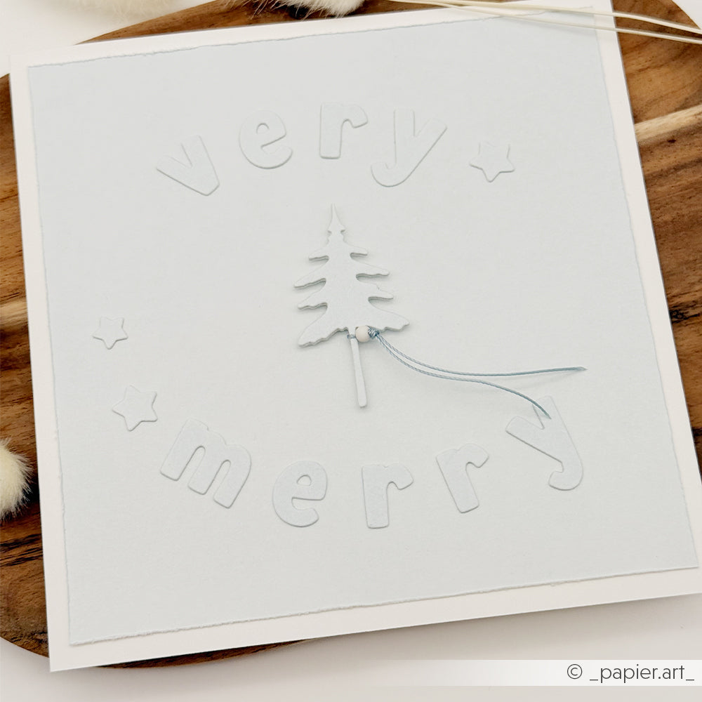Eine hellblaue, quadratische Weihnachtskarte mit dem geprägten Schriftzug „very merry“, der mit einer Motivstanze-Alphabet-Stanze gestaltet wurde, Sternen als Verzierung und einem zentralen Papierbaum aus Designpapieren.
